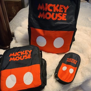 Könnte beinhalten: Ein schwarzer und roter Rucksack, eine passende Lunchtasche und ein Federmäppchen, alle mit dem Schriftzug MICKEY MOUSE in orangefarbenen Buchstaben und zwei weißen Kreisen auf dem roten Teil, die an die Hose der Figur erinnern.