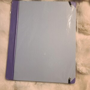 May include: A rectangular light blue folder with a purple spine and purple corner accents. The folder is enclosed in a clear plastic protective sleeve and features black elastic bands secured with silver-toned metal rivets at the corners.