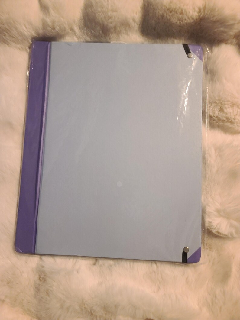 May include: A rectangular light blue folder with a purple spine and purple corner accents. The folder is enclosed in a clear plastic protective sleeve and features black elastic bands secured with silver-toned metal rivets at the corners.