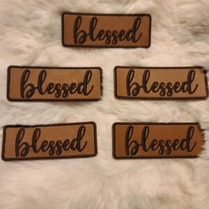 May include: Five rectangular tan patches with rounded corners, each featuring the word blessed embroidered in a dark brown cursive script. The patches have a dark brown stitched border and are arranged in a staggered formation on a white surface.