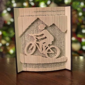 May include: A book art sculpture of a person riding a bicycle, made by folding the pages of a book. The sculpture is on a wooden table.