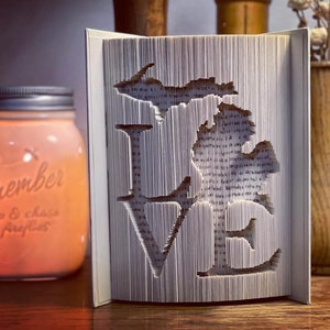 May include: A white book with the word "LOVE" cut out of the pages. The letters are formed by the pages of the book being folded and cut to create a 3D effect. The book is sitting on a wooden table.