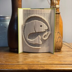 Könnte beinhalten: Eine Buchkunstskulptur eines Fisches, die aus den Seiten eines Buches gefertigt ist. Die Skulptur steht auf einem Holztisch mit zwei Holzvasen auf jeder Seite.