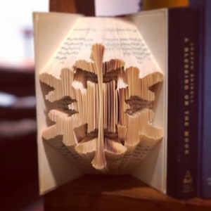 Könnte beinhalten: Ein Buch mit Seiten, die zu einem dreidimensionalen Schneeflocken-Design gefaltet sind. Das Buch ist geöffnet und die Schneeflocke ist in der Mitte des Bildes sichtbar. Das Buch liegt auf einer Holzoberfläche.