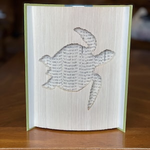May include: A book with pages folded and cut to create a three-dimensional sea turtle sculpture. The book is standing upright on a wooden surface.