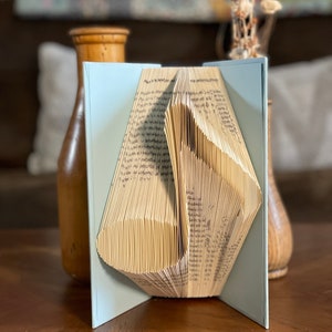 May include: A book folded into a three-dimensional shape, resembling a vase. The book is light blue with a cream-coloured interior. The book is sitting on a wooden surface.