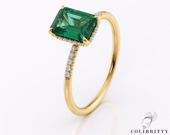 May include: A gold ring with a large emerald green gemstone surrounded by a halo of smaller diamonds. The ring has a thin band with a row of diamonds along the side.