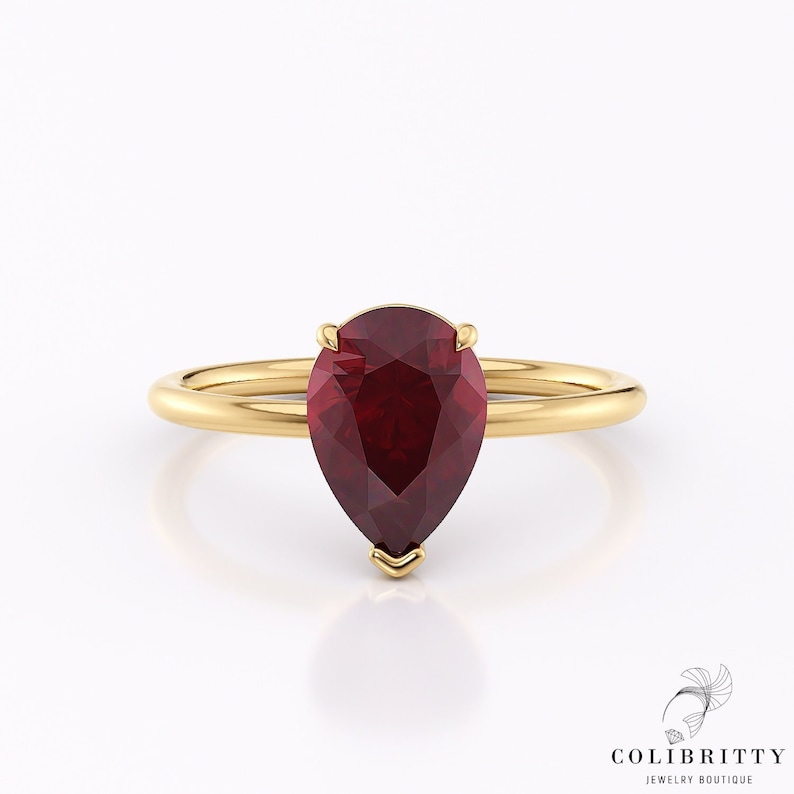 May include: A pear-shaped ruby gemstone set in a gold ring. The ring is simple and elegant, with a smooth band and a delicate setting for the ruby.