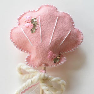 May include: A pink felt seashell wand with white stitching and decorative accents. The wand features a white ribbon bow and is embellished with pink beads and sequins. A whimsical accessory for imaginative play.