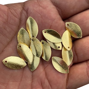 Puede incluir: Una colección de dijes metálicos dorados en forma de hoja con superficie texturizada. Cada dije tiene un pequeño agujero en la parte superior para ensartar. Los dijes se muestran en la mano de una persona, mostrando su tamaño y detalle. Ideal para la creación de joyas.