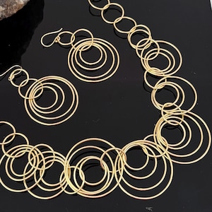 May include: A gold-toned jewelry set consisting of a necklace and a pair of earrings. The necklace features a series of interconnected circular rings of varying sizes. The matching earrings are composed of smaller clusters of linked gold circles.
