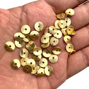 May include: A collection of small, gold-coloured, disc-shaped beads with a central hole. The beads have a textured surface. Suitable for jewellery making and crafting. Each bead is approximately 1 cm in diameter.