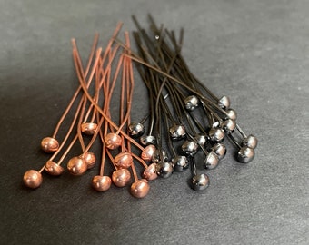 May include: A collection of thin metal head pins with small spherical ends, arranged in two distinct groups. One group features a warm copper finish, while the other group consists of pins with a dark, gunmetal grey metallic finish.