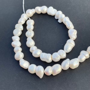 Könnte beinhalten: Eine Strähne weißer Barockperlen mit unregelmäßigen Formen und Größen, auf einem weißen Faden aufgereiht. Die Perlen sind in einem lockeren, fließenden Muster angeordnet.
