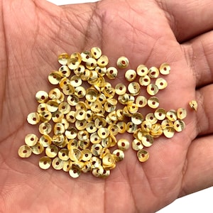 Może przedstawiać: Duża sterta złotych metalowych cekinów rozrzuconych na dłoni. Ceki są okrągłe z małym otworem w środku.