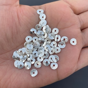 Peut inclure: Un tas de perles en métal de couleur argent, chacune ayant une forme plate et circulaire avec un centre légèrement surélevé. Les perles sont disposées de manière aléatoire sur une main.