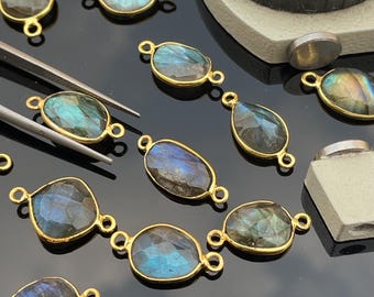 May include: A collection of faceted labradorite gemstones set in gold-toned metal frames with two loops on opposite sides. The stones display iridescent blue and gray flashes. A metal caliper is visible in the corner for scale measurement.