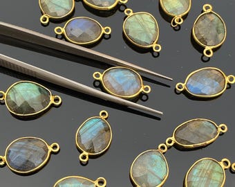 May include: A collection of faceted, teardrop-shaped labradorite gemstones set in gold-toned metal bezels with two small loops on opposite ends. Each stone displays iridescent blue and green flashes, and a pair of metal tweezers is visible.
