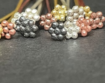 May include: A collection of hair pins featuring decorative tops shaped like small clusters of spheres. The tops are finished in metallic tones of silver, gold, copper, and dark charcoal gray, each with a textured, granular surface appearance.
