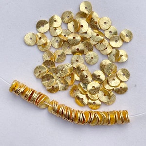 May include: A large quantity of gold-coloured metal round disc beads. The beads are arranged in a pile and a string of beads is laid out on a white surface.