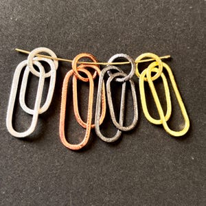 May include: Four sets of two interlocking oval-shaped metal links arranged in a horizontal row. The sets are finished in silver, copper, dark gray, and gold tones, each featuring a textured surface and suspended on a thin, curved gold-toned wire.