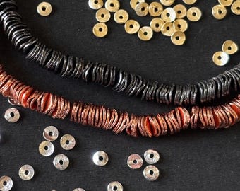 Puede incluir: Primer plano de componentes de joyería, incluyendo dos hilos de cuentas en forma de disco en colores negro y cobre. Dispersas, pequeñas cuentas metálicas doradas y plateadas en forma de disco, todo sobre una superficie negra. Las cuentas tienen una apariencia texturizada.