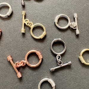 Puede incluir: Una colección de cierres de palanca para joyería, que incluye anillos circulares y componentes de barra en T. El conjunto presenta varios acabados metálicos como oro, plata, oro rosa y metal plomo, cada uno con detalles estriados.