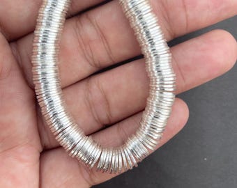 Puede incluir: Un hilo de pequeñas cuentas circulares y planas de color plateado dispuestas en un bucle continuo. Cada cuenta presenta una forma delgada de disco con un acabado metálico, creando una apariencia uniforme y reflectante a lo largo del hilo.