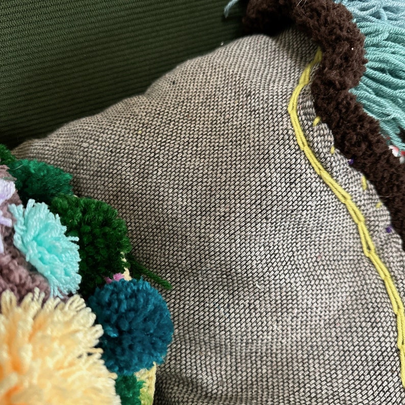 May include: A gray throw pillow with a yellow embroidered design and a colorful pom-pom trim. The pom-poms are in shades of green, blue, pink, and yellow.