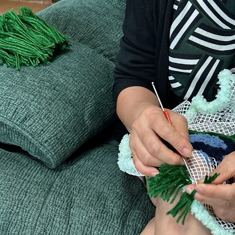 May include: A person is working on a green and blue rug with a white mesh backing. They are using a crochet hook to attach green yarn to the mesh. The rug is being made with a variety of colors and textures.