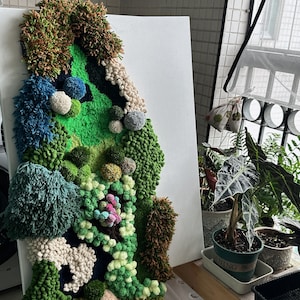 May include: A green, blue, and white yarn wall art piece depicting a landscape scene. The piece is made of various textures and colors of yarn and is mounted on a white canvas.