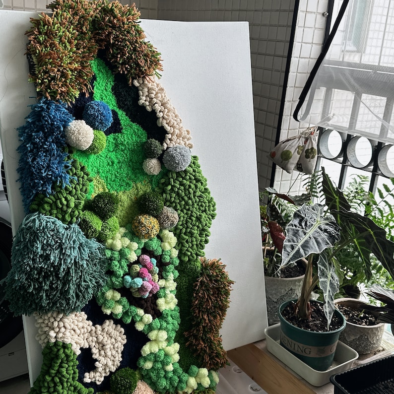 May include: A green, blue, and white yarn wall art piece depicting a landscape scene. The piece is made of various textures and colors of yarn and is mounted on a white canvas.
