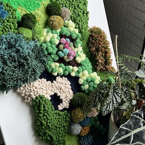 May include: A close-up of a wall hanging made of yarn in various shades of green, white, and brown. The yarn is arranged in a textured, abstract pattern, resembling a landscape or garden.