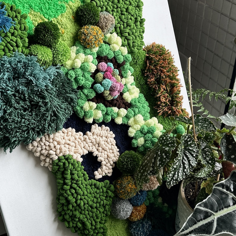 May include: A close-up of a wall hanging made of yarn in various shades of green, white, and brown. The yarn is arranged in a textured, abstract pattern, resembling a landscape or garden.