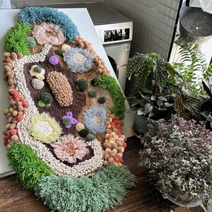 Könnte beinhalten: Ein bunter, handgefertigter Teppich mit verschiedenen Texturen und Formen. Der Teppich ist aus Garn und Pompons in den Farben Grün, Blau, Rosa und Braun gefertigt.