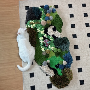 May include: A white cat sits next to a green, brown, and blue rug with a textured, fluffy surface. The rug is shaped like a cloud and has a variety of different textures.