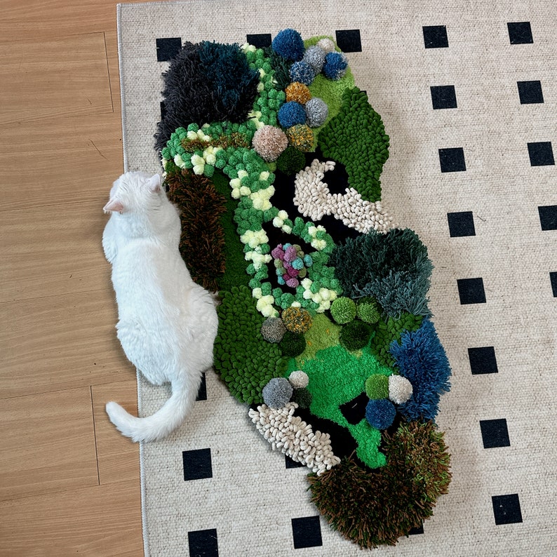 May include: A white cat sits next to a green, brown, and blue rug with a textured, fluffy surface. The rug is shaped like a cloud and has a variety of different textures.