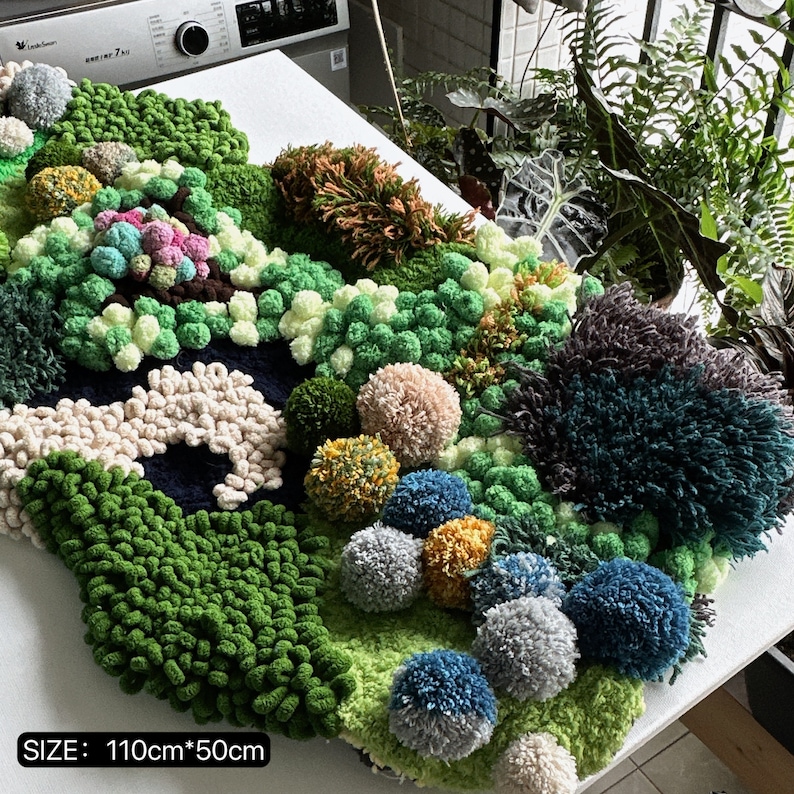 May include: A green, blue, and brown wall hanging made of yarn pom-poms and fuzzy fabric. The wall hanging is 110 cm by 50 cm.
