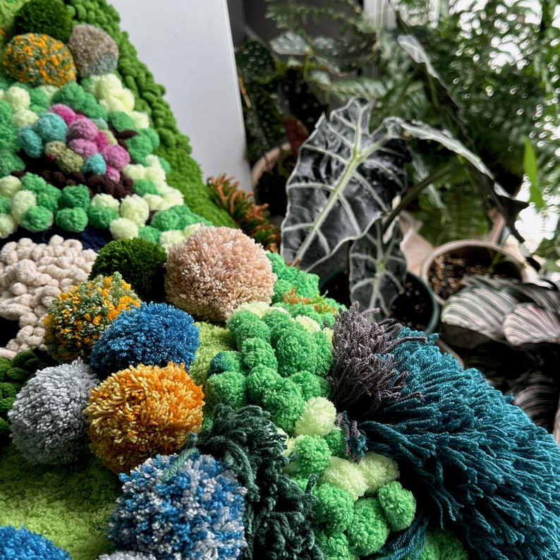 May include: A close-up of a green, blue, and yellow yarn wall hanging with a variety of textures. The wall hanging is made of pom-poms, yarn, and other materials.