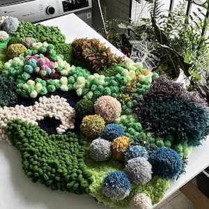 May include: A green, blue, and white wall hanging made of yarn pom-poms and fuzzy fabric. The wall hanging is shaped like a cloud and has a variety of textures.