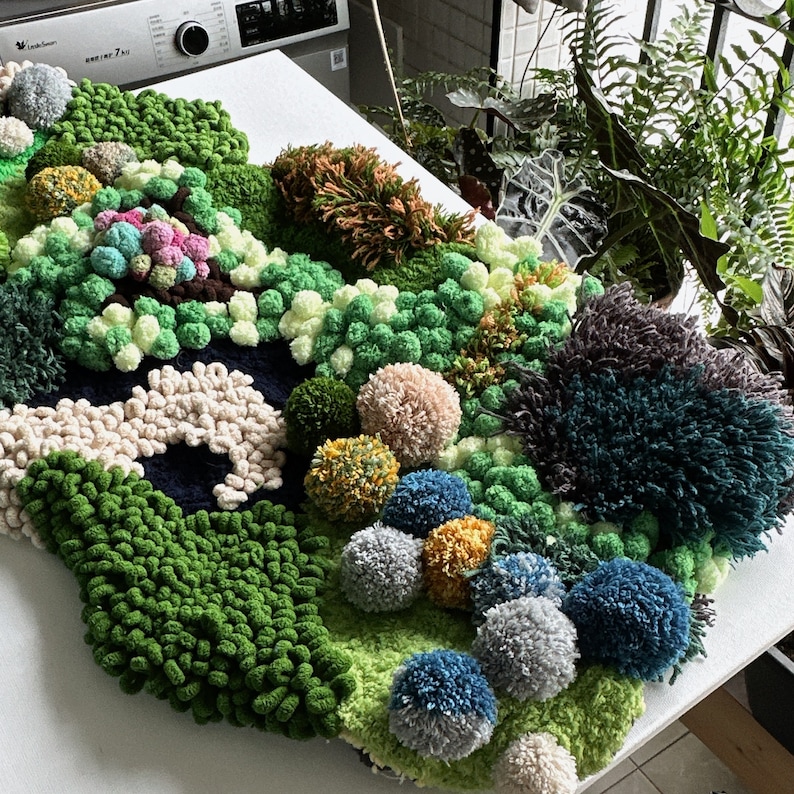 May include: A green, blue, and white wall hanging made of yarn pom-poms and fuzzy fabric. The wall hanging is shaped like a cloud and has a variety of textures.