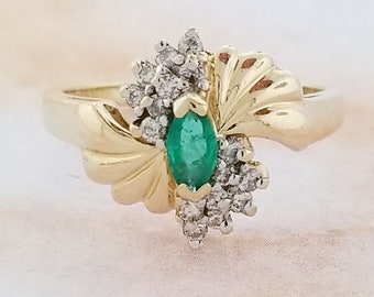 May include: A gold ring with a green emerald gemstone surrounded by small diamonds. The ring has a unique design with two gold leaves on either side of the emerald.