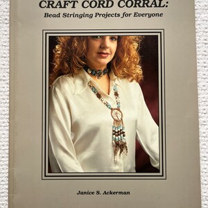 May include: Book cover for "Craft Cord Corral: Bead Stringing Projects for Everyone" by Janice S. Ackerman. The cover features a woman wearing a beaded necklace with turquoise and brown beads.