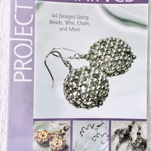 May include: Book cover with the title "EARRINGS PROJECT" and the subtitle "44 Designs Using Beads, Wire, Chain, and More". The cover features three different earring designs, including a pair of silver bead earrings, a pair of earrings with purple and gold beads, and a pair of silver wire earrings.