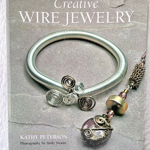 May include: A book titled "Creative Wire Jewelry" by Kathy Peterson with photography by Andy Newitt. The cover features a silver wire bracelet and a pendant with a brown and gold stone wrapped in wire.