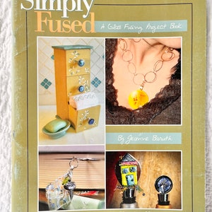 May include: Book cover with the title "Simply Fused: A Glass Fusing Project Book" by Jeanne Baruth. The cover features four images of fused glass projects, including a small dresser, a necklace, a bottle stopper, and a house-shaped bottle stopper.