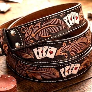 May include: A brown leather belt featuring an embossed feather pattern and a repeating graphic of four playing cards, including an ace of hearts. The belt has silver-toned metal rivets and a dark, weathered finish on the leather surface.