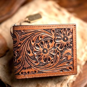 May include: A brown leather wallet with a floral embossed design. The wallet features a detailed pattern of flowers and leaves, with a tag attached. The wallet is rectangular and has a stitched border.