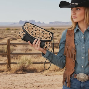 May include: Black and tan leather clutch bag with floral tooling. The bag features a wrist strap and a snap closure. The design includes a black base with tan floral detailing. The background shows a desert landscape.