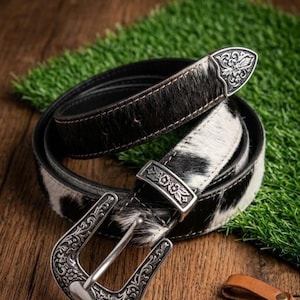 Puede incluir: Un cinturón de piel de vaca en blanco y negro con hebilla y punta plateadas. El cinturón tiene un diseño floral decorativo en la hebilla y la punta. El cinturón está enrollado sobre una superficie de madera.